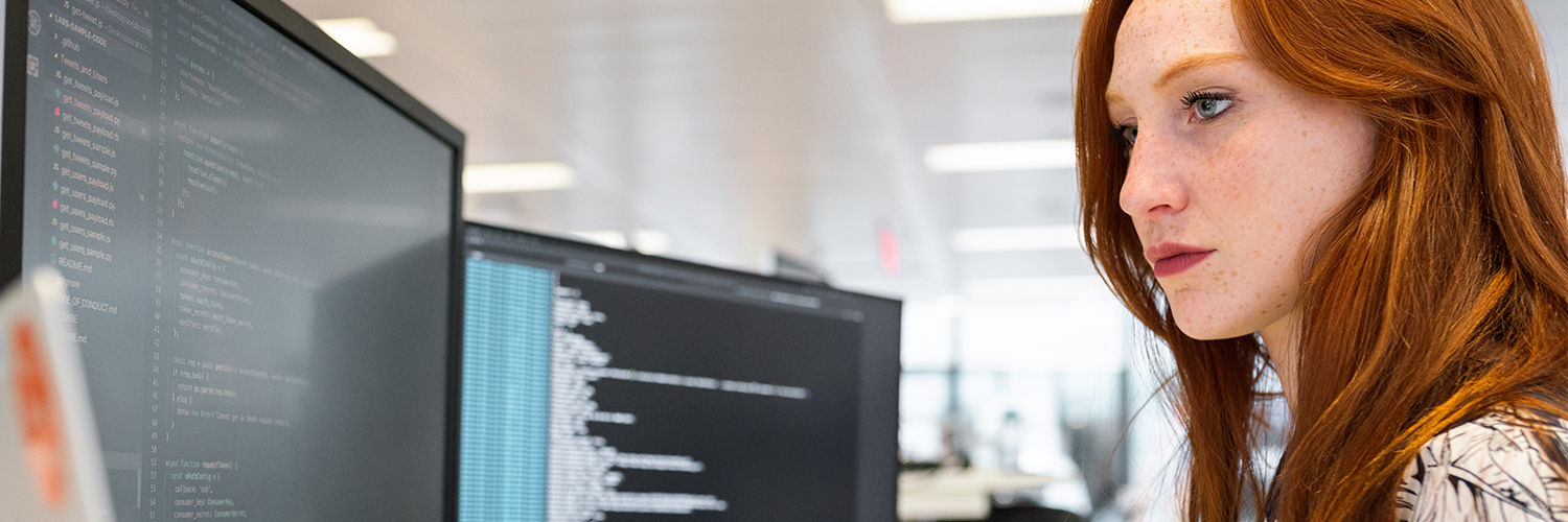 Female computer scientist looking at two monitors with coding on the screen