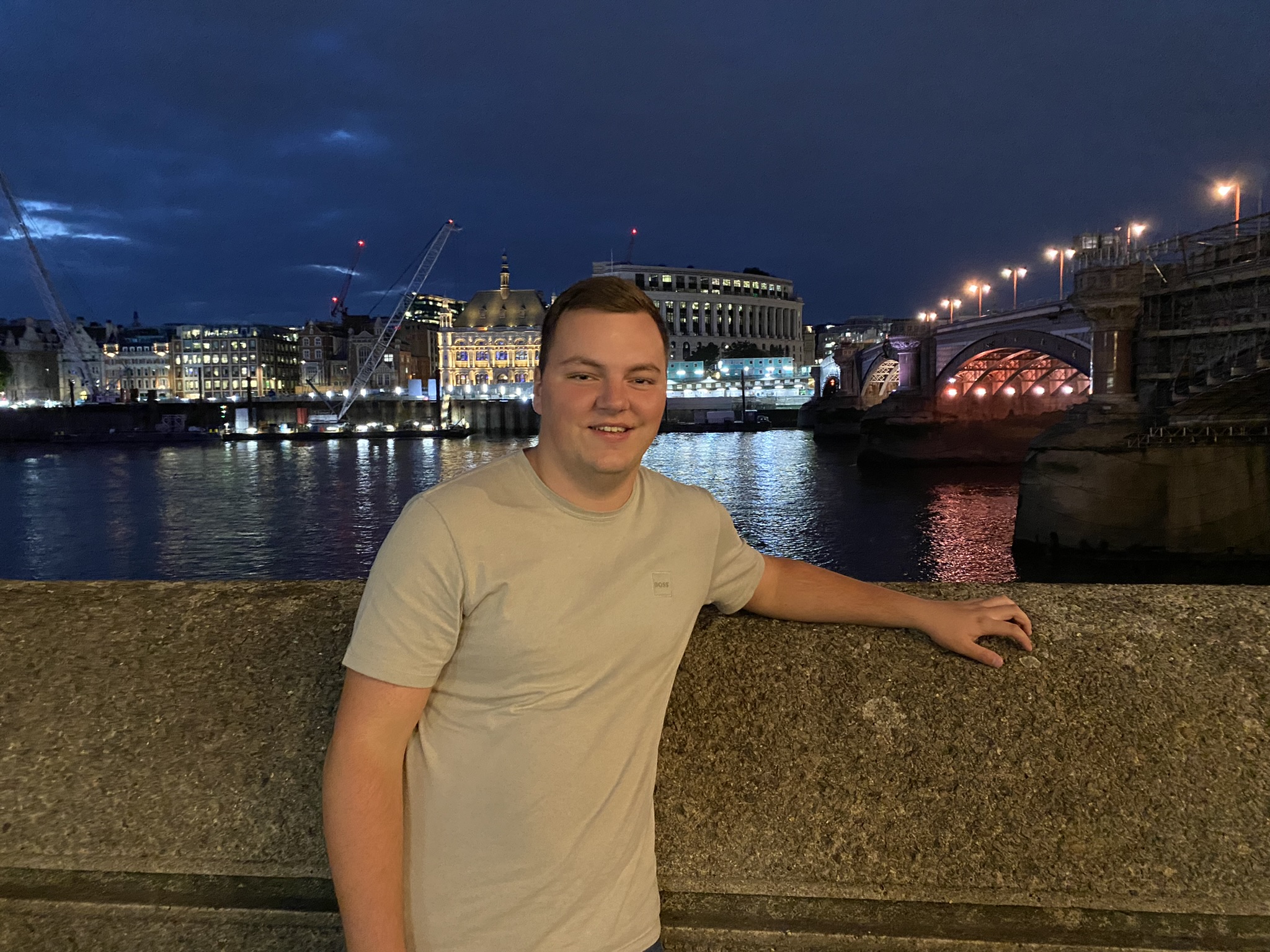 Salford student Lloyd Townsend stood in front of a stone wall at night with river and lit up building behind him.