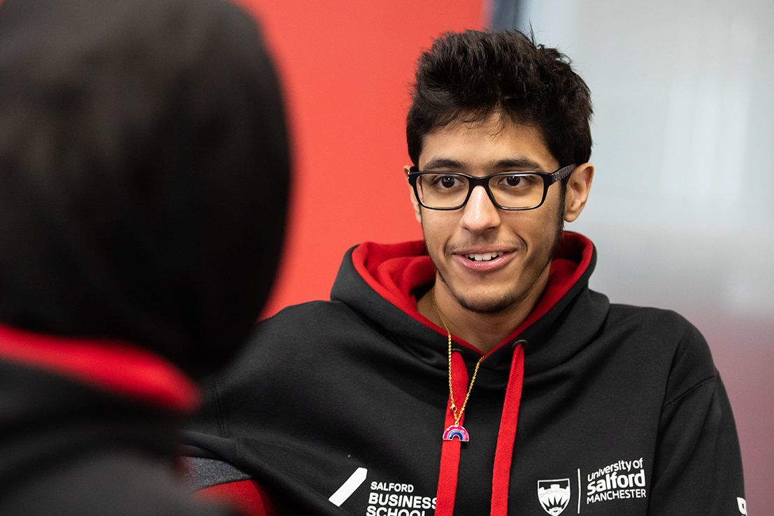 A student wearing a black and red university hoodie, smiling and talking to another person, with a blurred red and grey background.