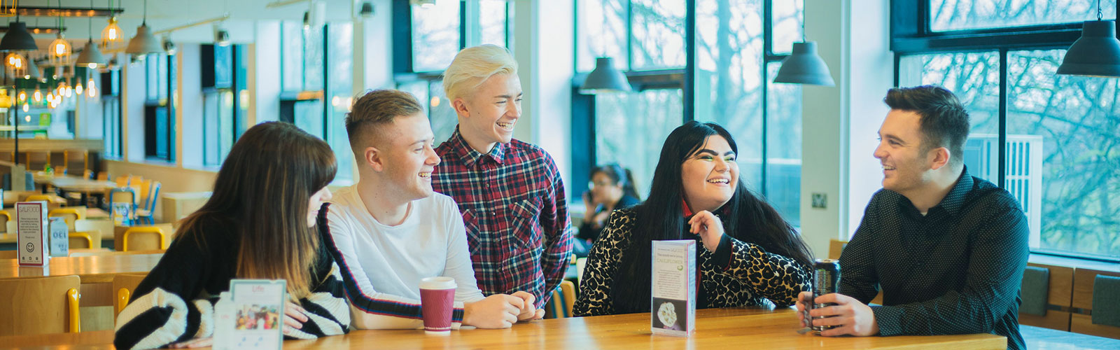 Five students socialising around a table in Maxwell Café, University of Salford