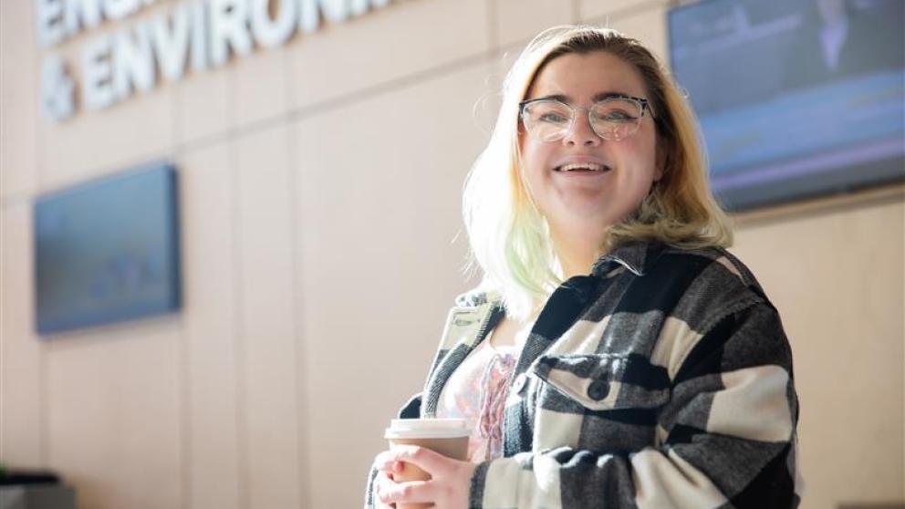 Student in black and white check shirt stood holding coffee cup smiling