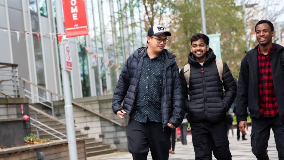 Three male students walking across our Peel Park campus in front of our New Adelphi building