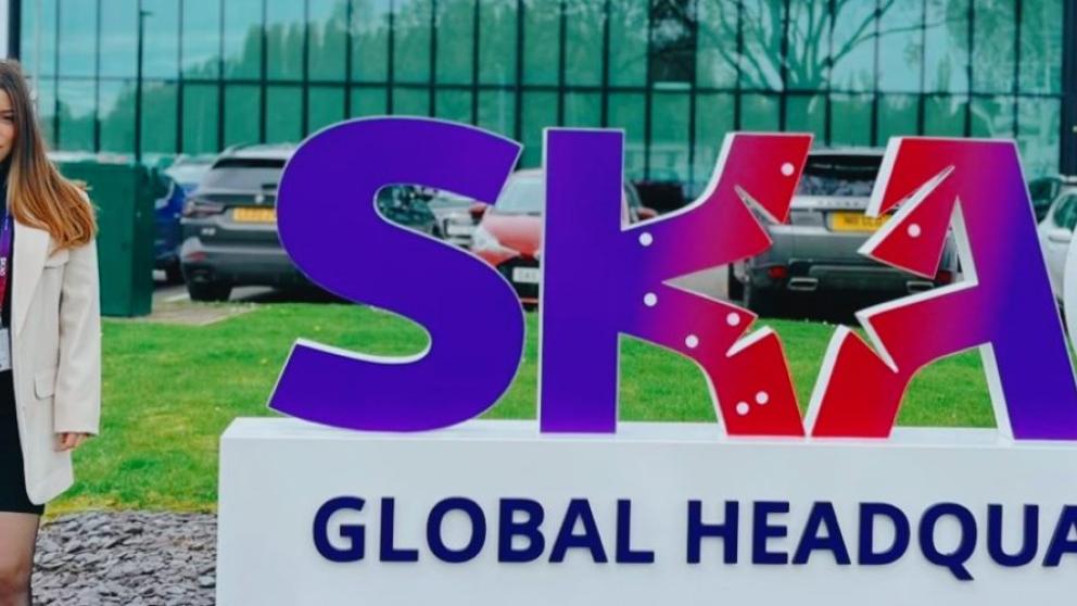 A female student in a blazer outside a sign for SKAO