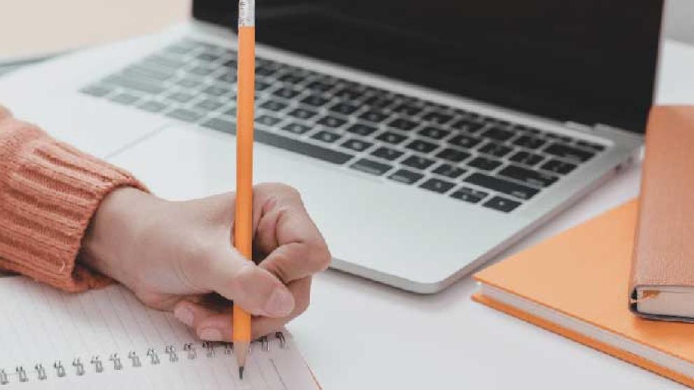 Person writing in a notebook with a pencil next to an open laptop and closed notebooks