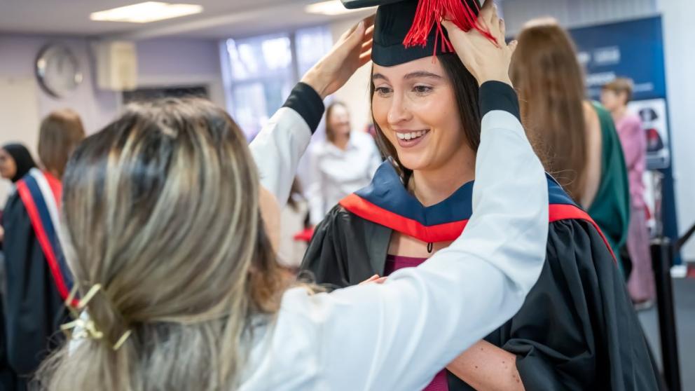 Student gets graduation cap placed on head by staff member in robing area