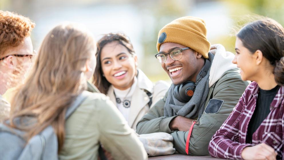 Group of male and female students sat around a table chatting outside